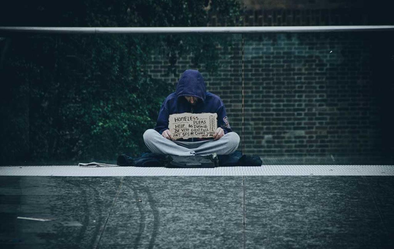 A beggar sits cross legged against a glass railing holding a sign.