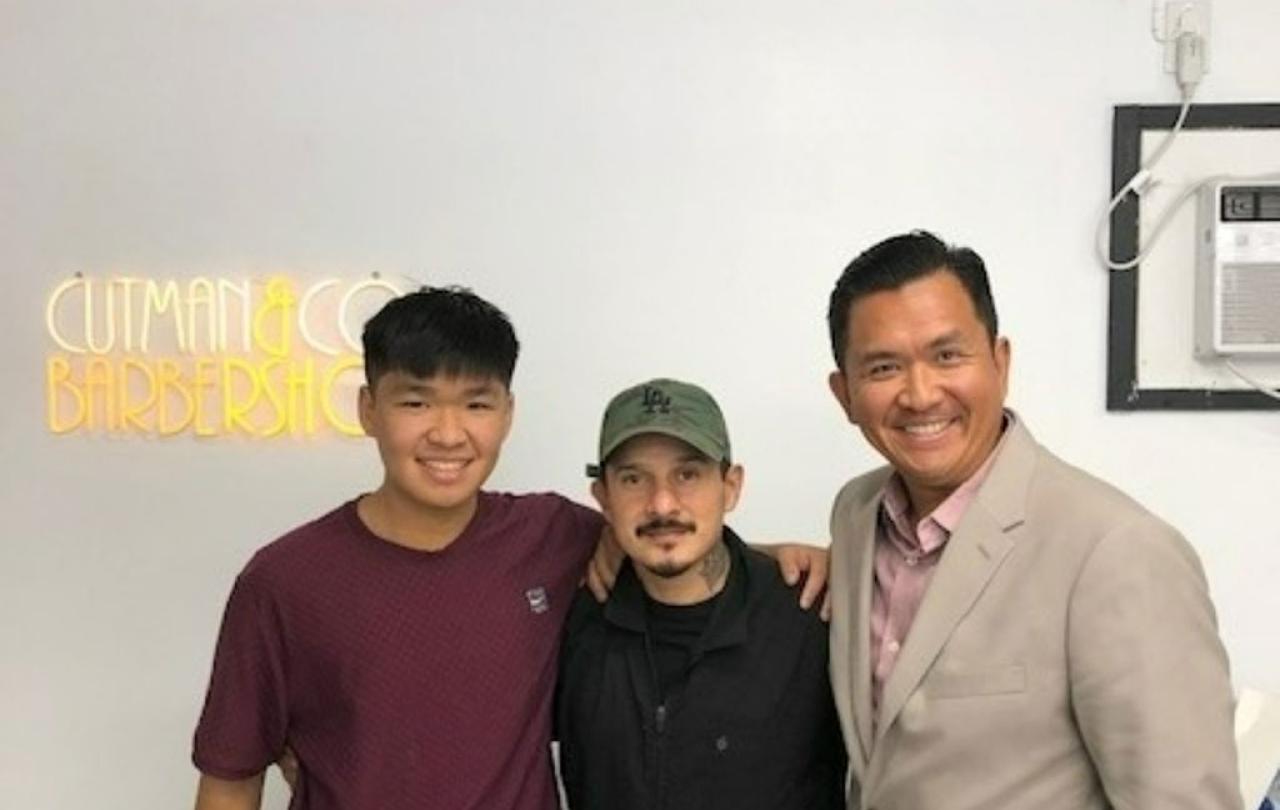 A barber stands between two clients, a father and son, a neon sign shines behind.