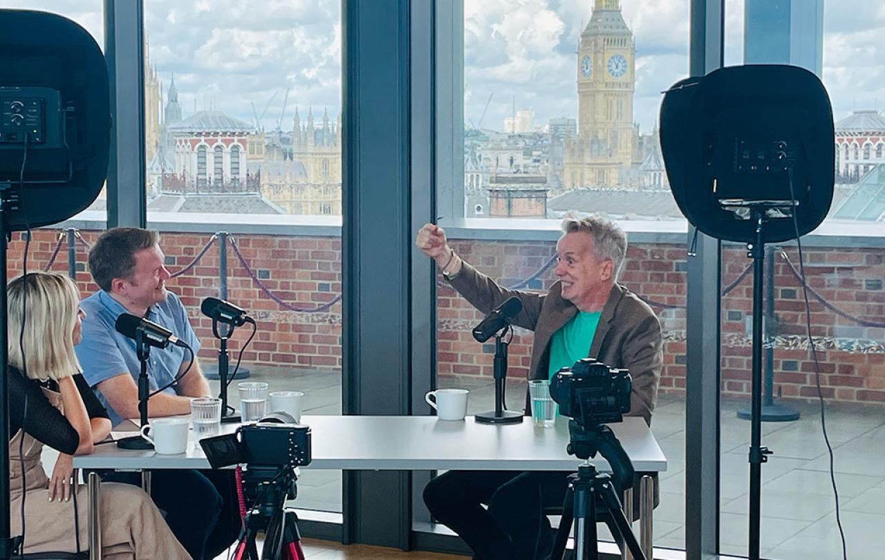 At a podcast recording in a room overlooking Big Ben, A man holds his hand up in gesture while speaking