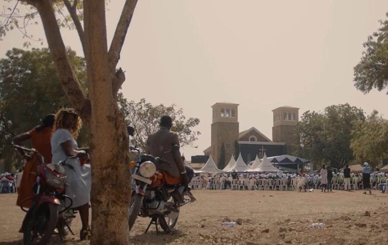 Outside a church a congregation waits seated while an onlooker rests on a motorcycle by a tree.