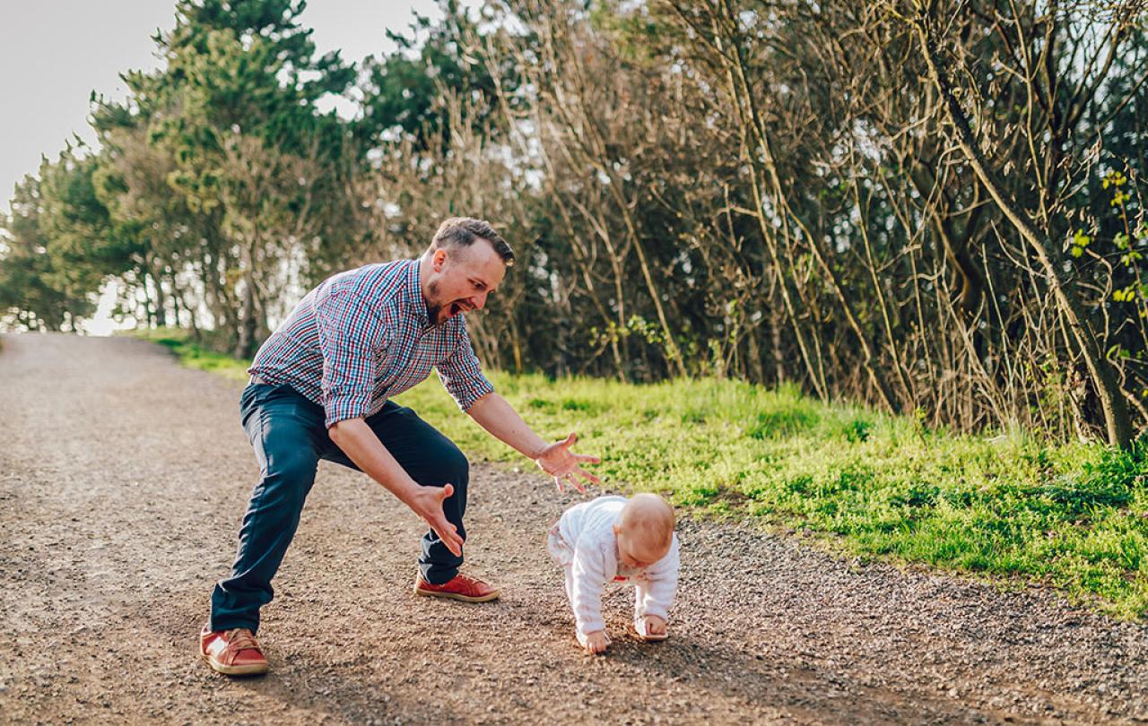 A dad hovers with open arms ready to catch a baby taking first steps