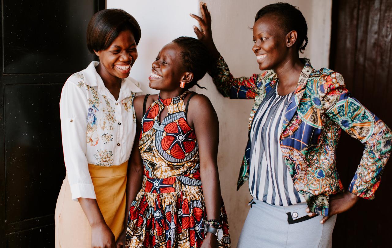 Three women stand, two lean into each other sharing a joke, while the other laughs too.