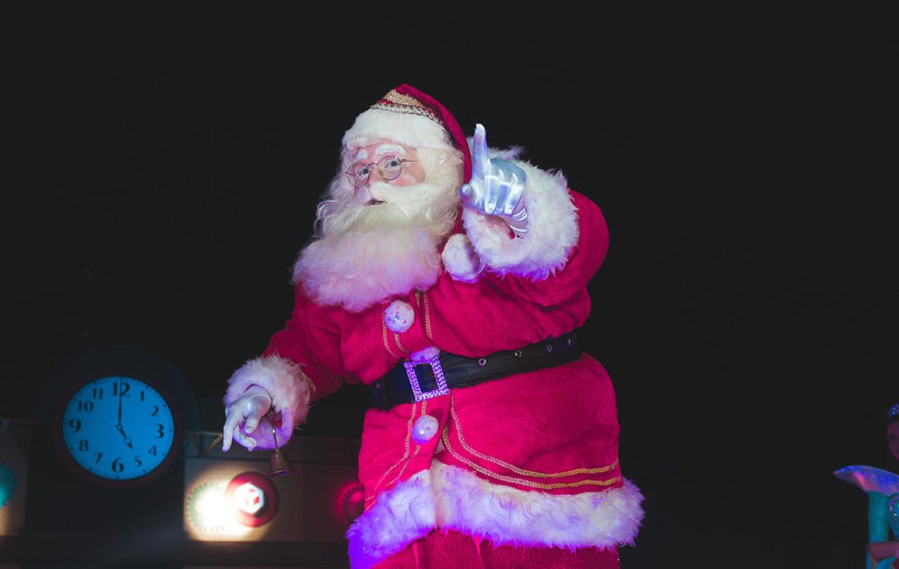 A Santa tiptoes in a darkened room carrying a lamp and holding up a finger in a gesture of silencing