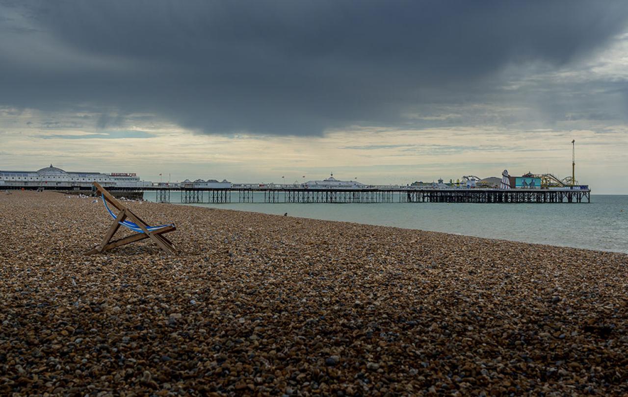 A moody sky overshadows a shingle beach on which a lone empty deckchair stands. A pier with funfair is in the middle distance.