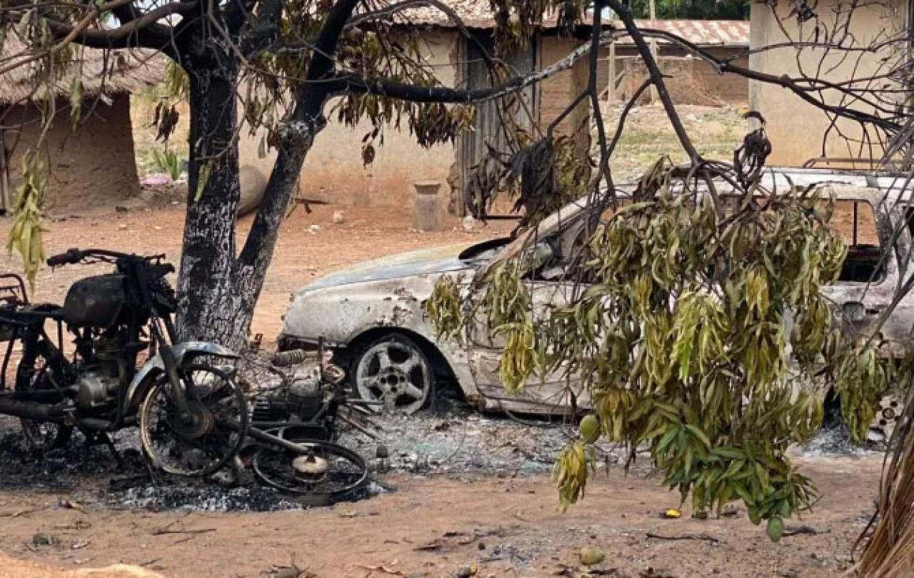 A burnt out motor cycle and car stand amid charred debris in a dusty compound.