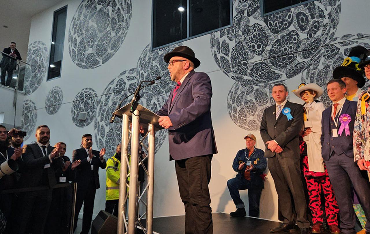 A winning candidate at an election address the audience from a lectern while the loosing candidates look on