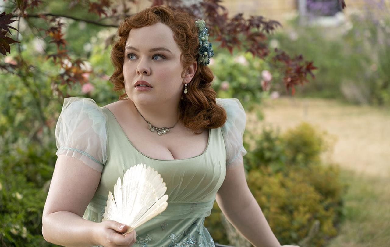A young lady in Regency dress, holds a fan while looking around a garden.