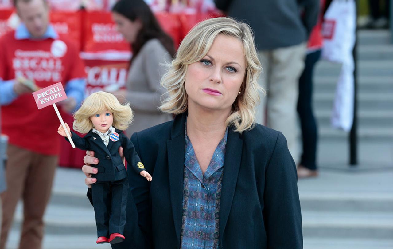 A deflated looking woman stands aside from a protest rally, holding a small doll of herself that reads Recall Knope'.