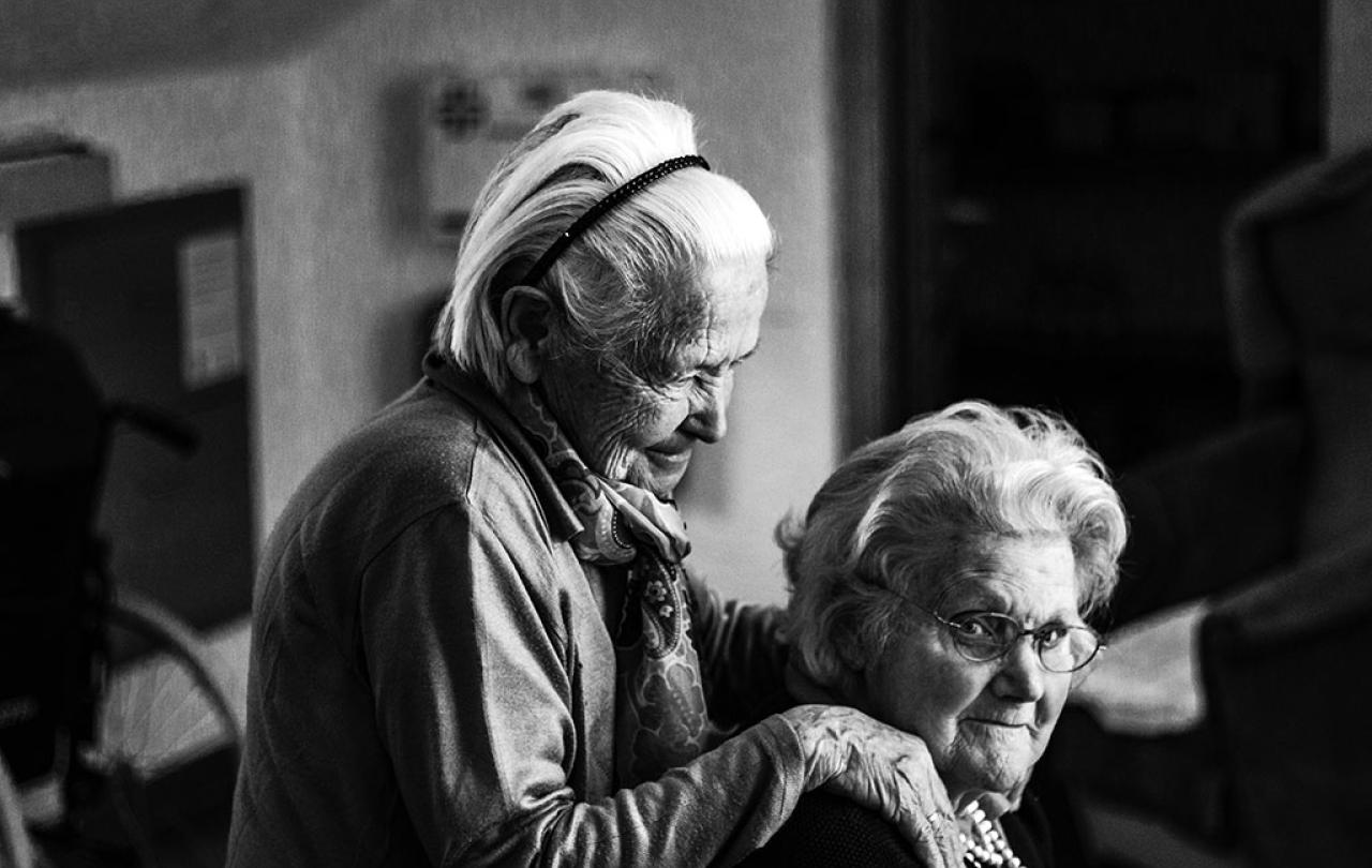 A elderly women in a care home stands and places her hands on the shoulders of a seated woman.