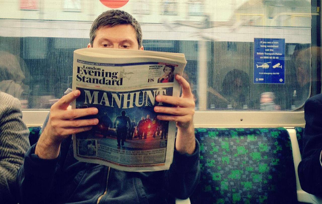 Seat on an underground train carraige, a passenger holds and reads a newspaper.