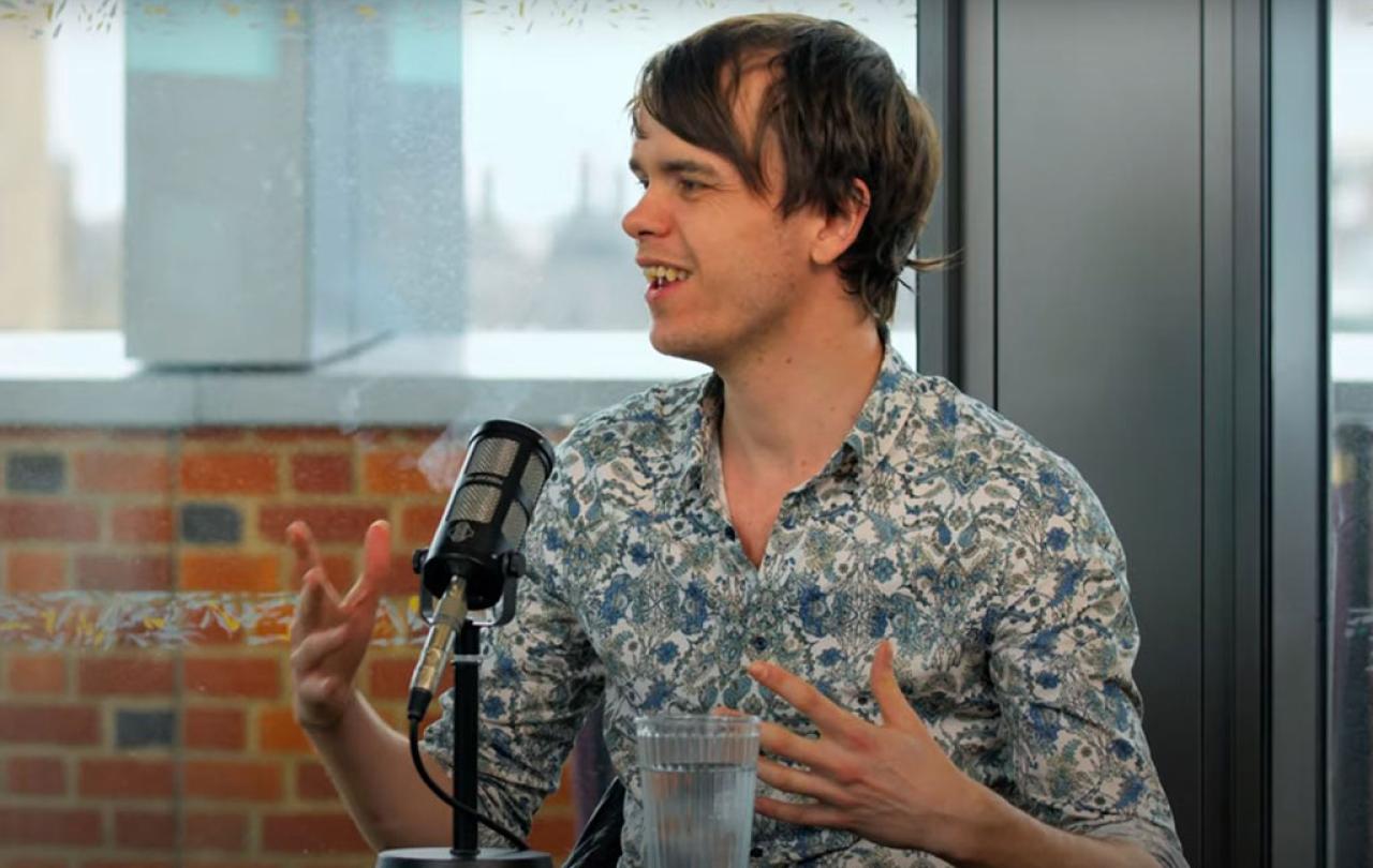 A podcast guest speaks in front of a mic and raises their hands to gesture.