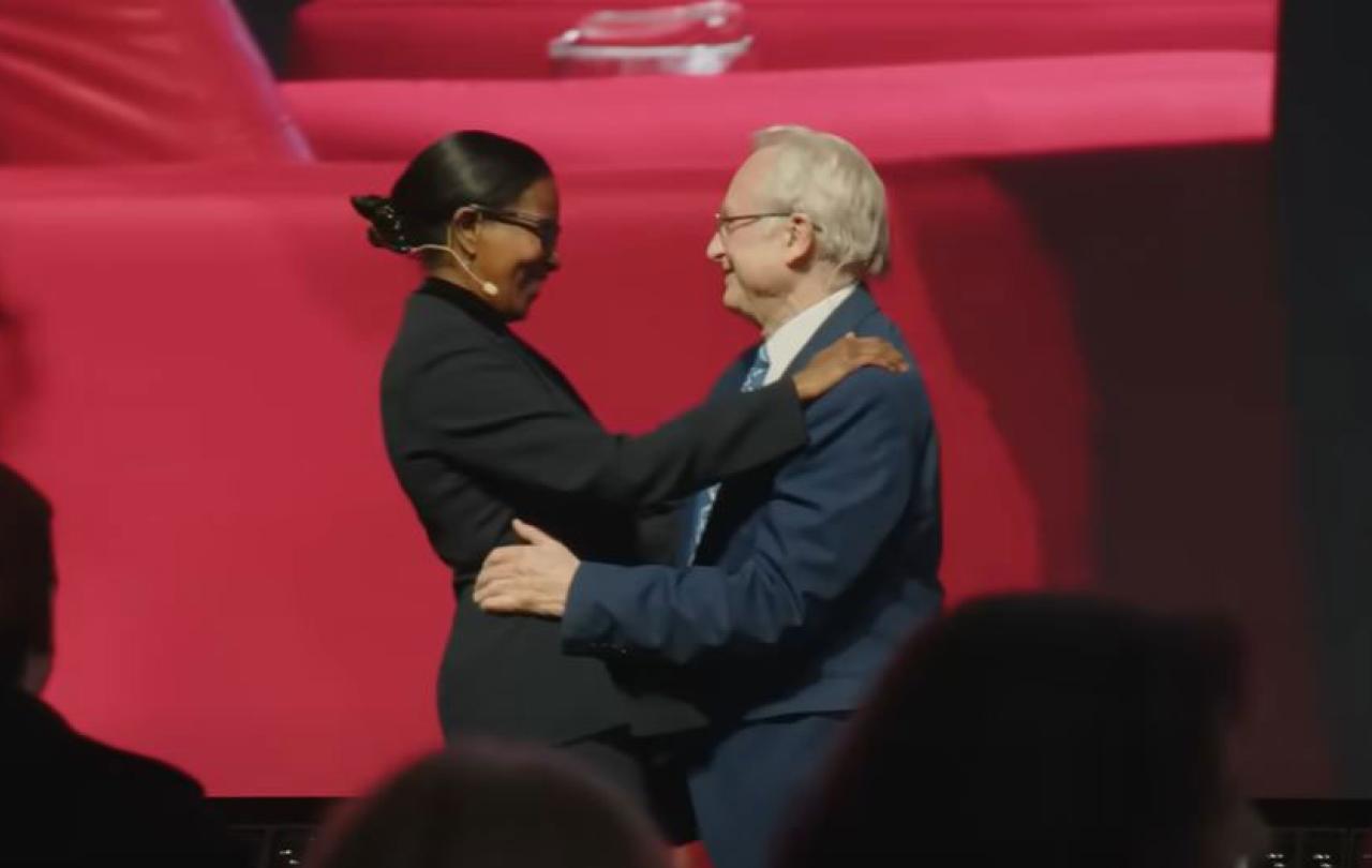 A man and woman speaker on a stage greet and embrace each other.