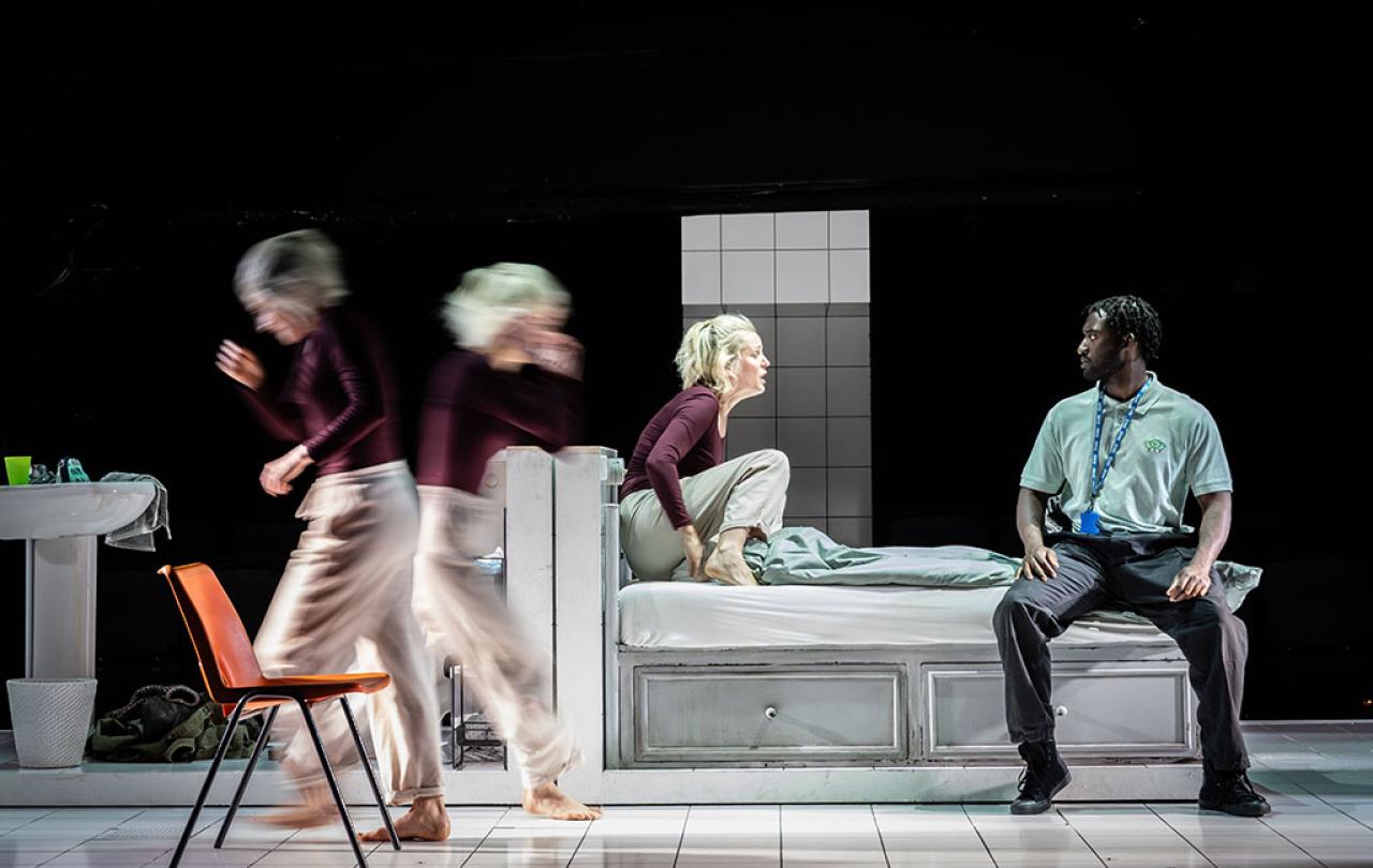 on a stage a woman kneels on a bed amid frantic action around her.