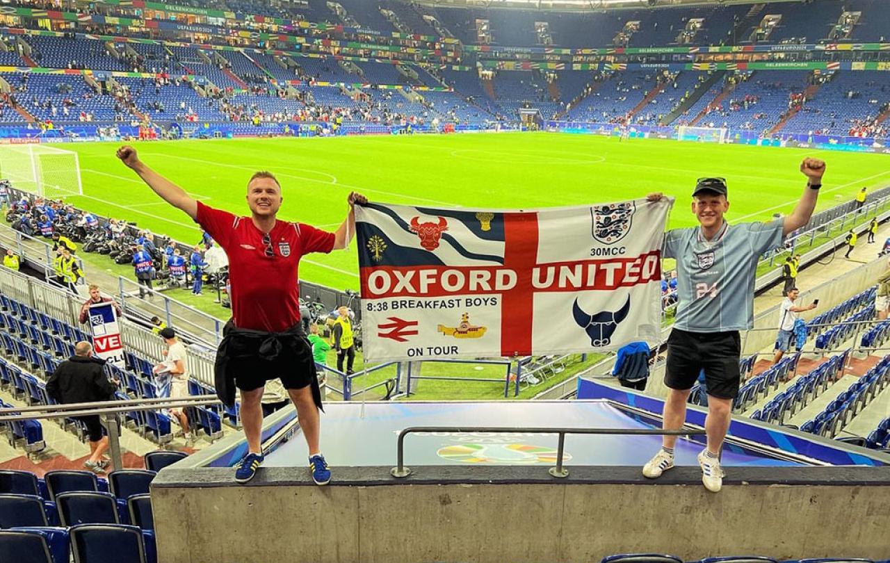 Two England fans stand in stadium seeting holding up their flag.