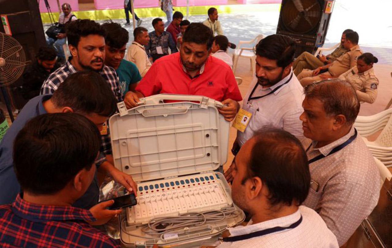 official surround an open case containing an electronic voting terminal and cables.