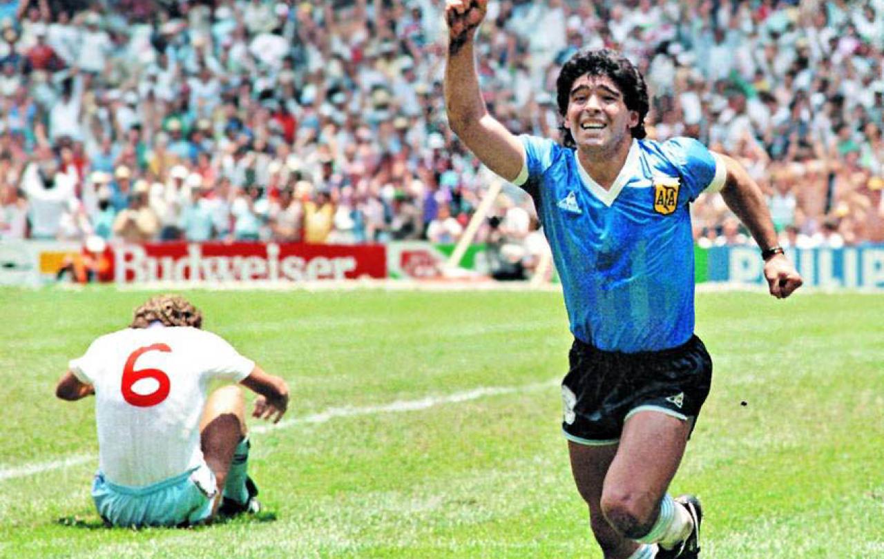 Maradona runs in celebration, holding a hand aloft as an England player sits dejected on the ground.