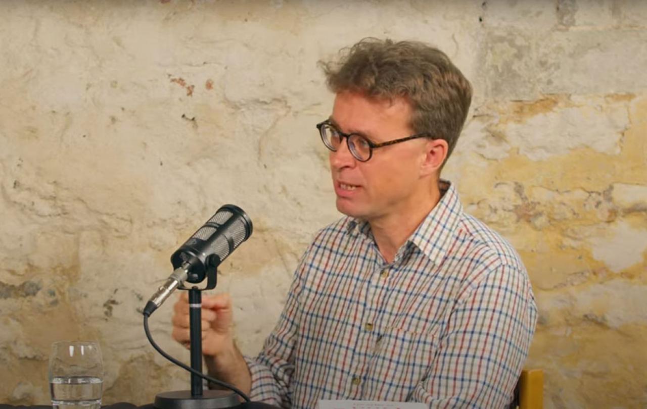 A podcast guest speaks in front of a mic and raises their hands to gesture.