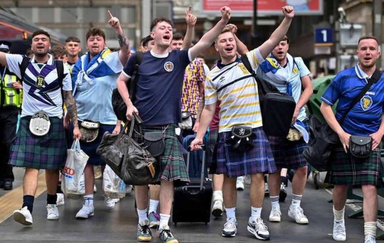 Scottish football fans wearing kilts march down a street singing and waving their arms alogt.
