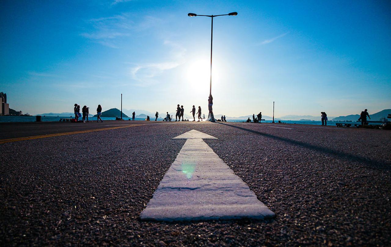 A white arrow on tarmac points towards a setting sun and people walking by a silhouetted lamp post.