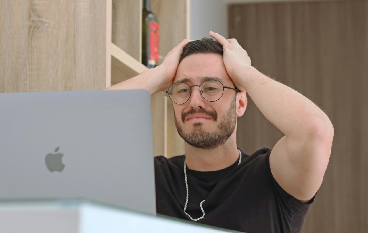 A man staring at a laptop grimmaces and holds his hands to his head.