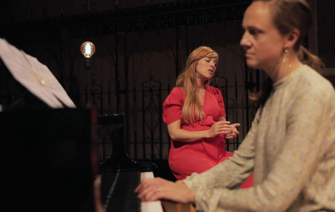 A pianist plays in the foreground and a seated singer gestures with eyes closed behind 