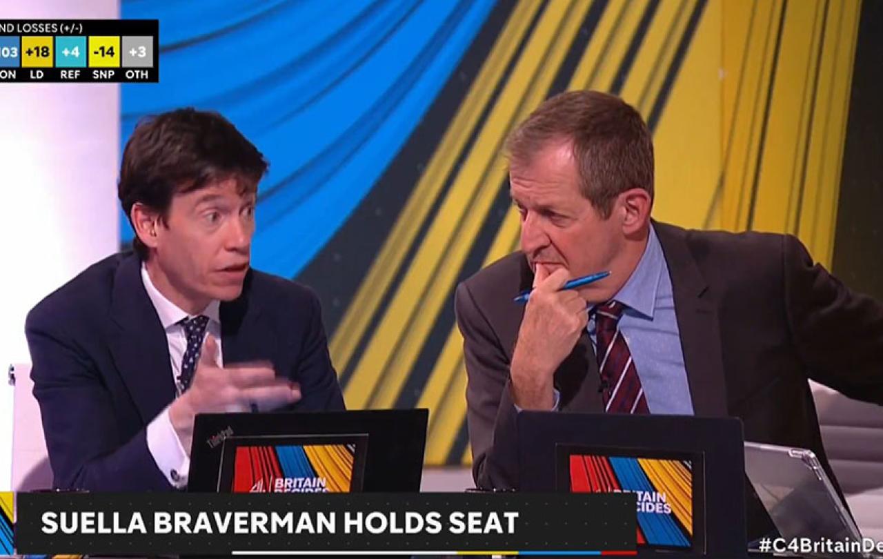 A poltiical pudit opines in a TV studio while his colleague leans in and listens.