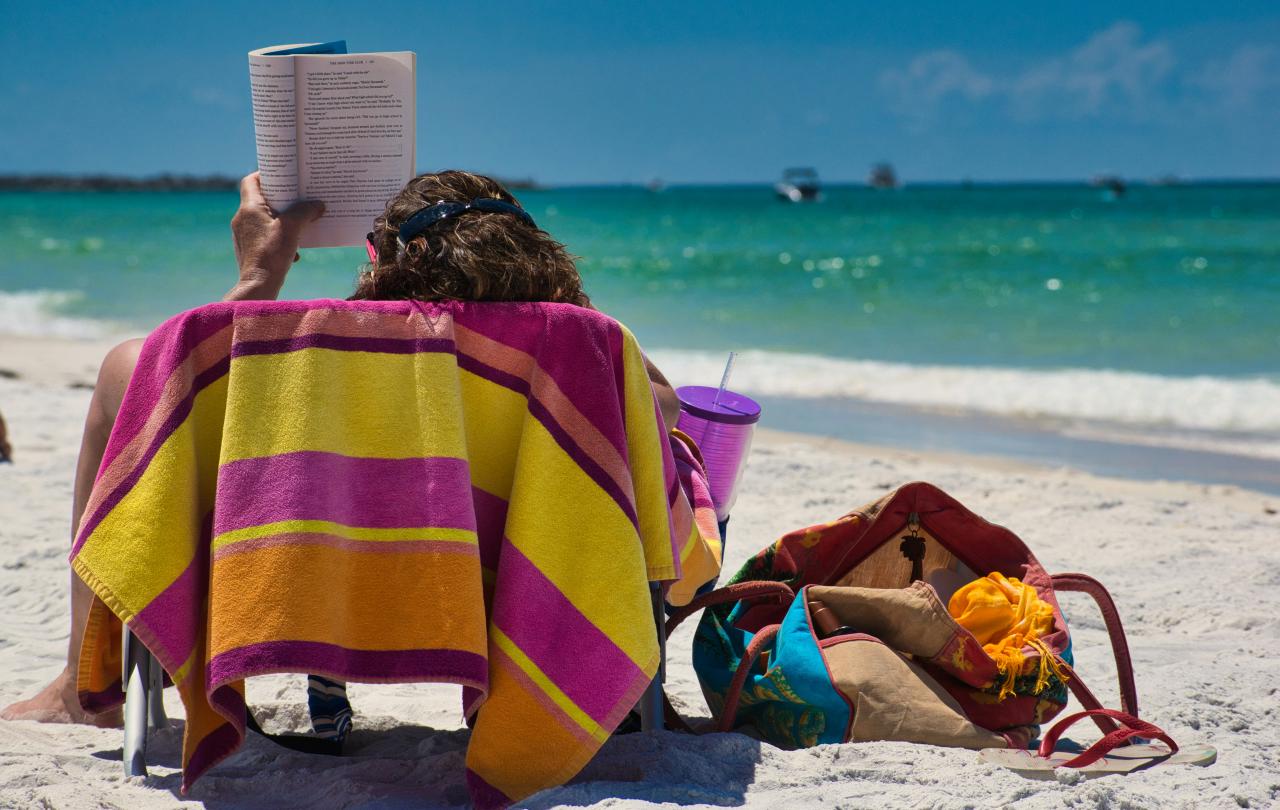 On a beach lounger someone holds a book aloft to read.