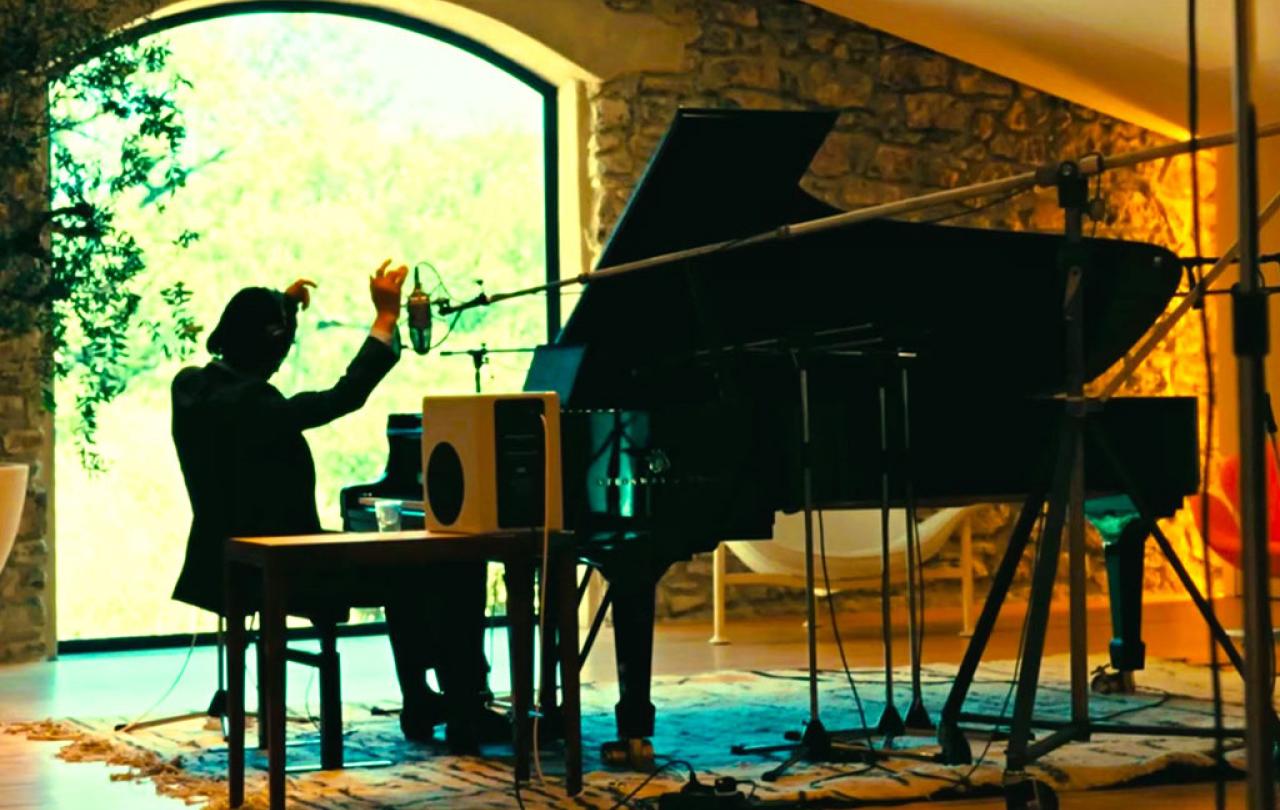 A pianist raises his arms while sitting at a grand piano amid recording equipment.