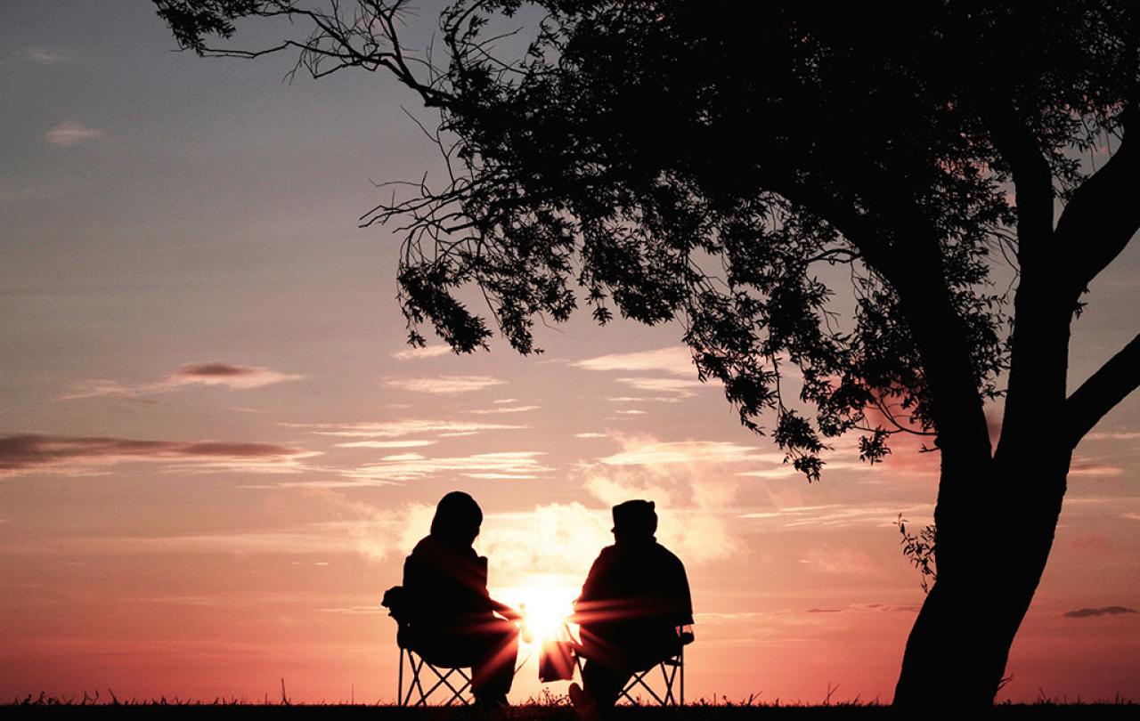 Under a tree, backlit by a sun set, two people sit in chairs outside and talk.