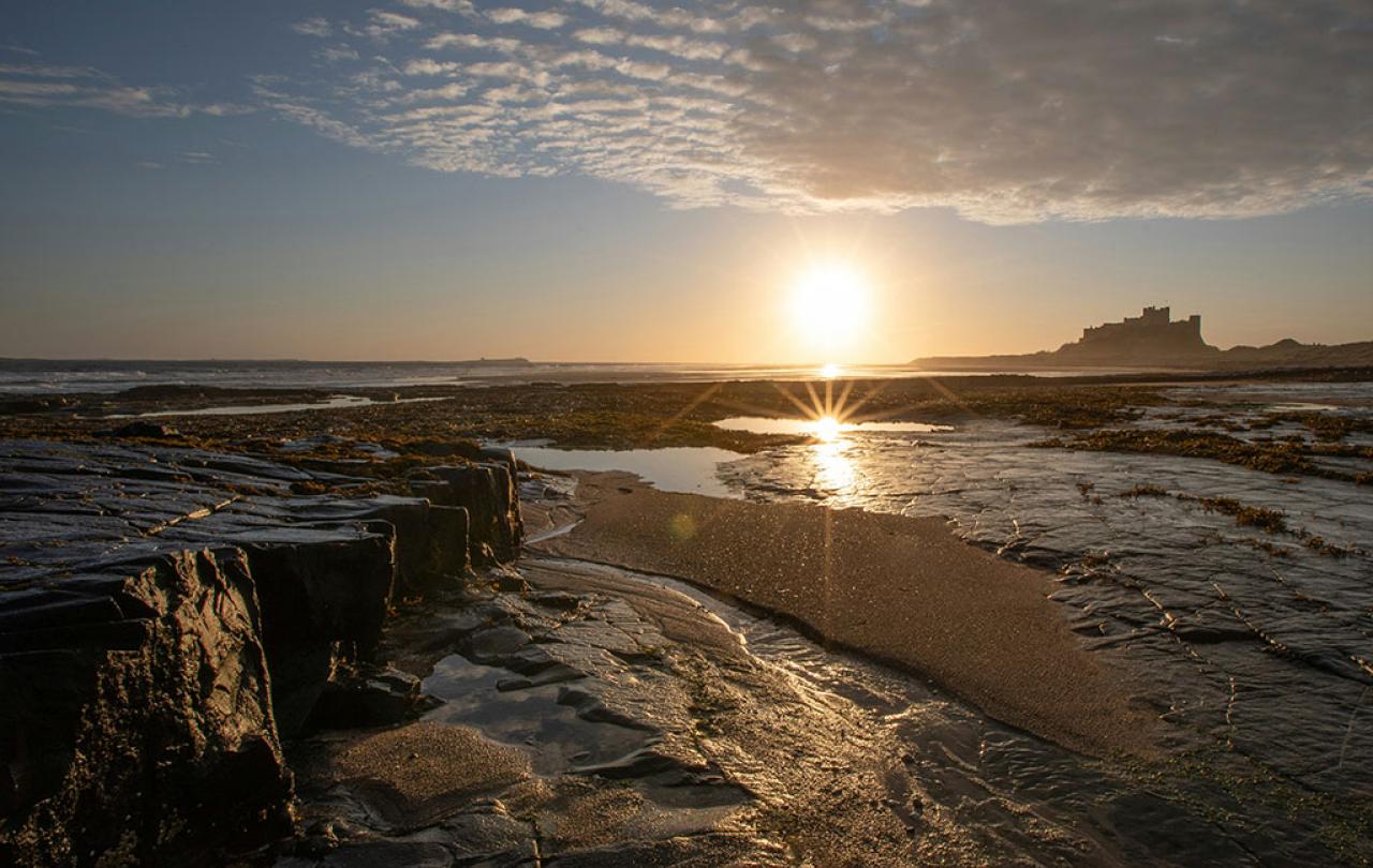 A rockpool on a beach reflects the sun, a castle stands beyond the sand dunes