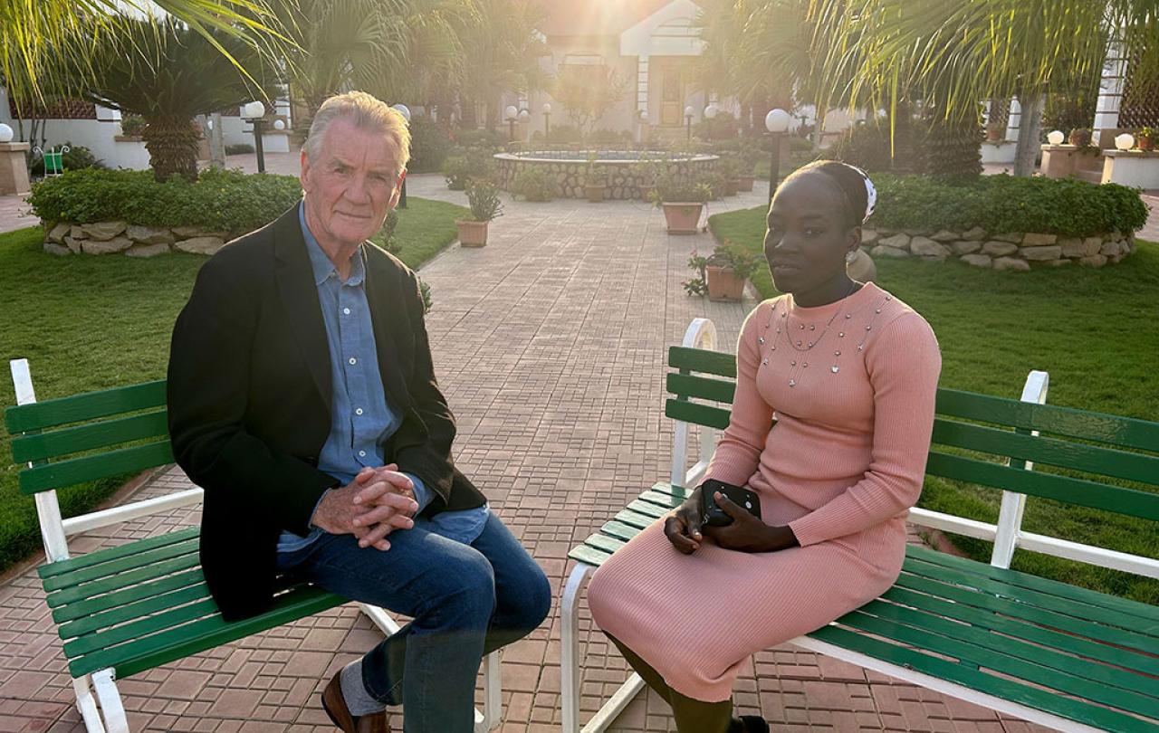 A TV interviewer sits across from a young woman in an outdoor setting