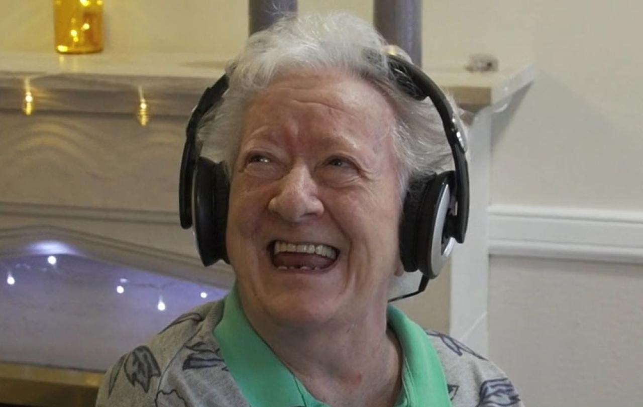 An elderly woman wearing headphone looks up and to the side with a big smile.