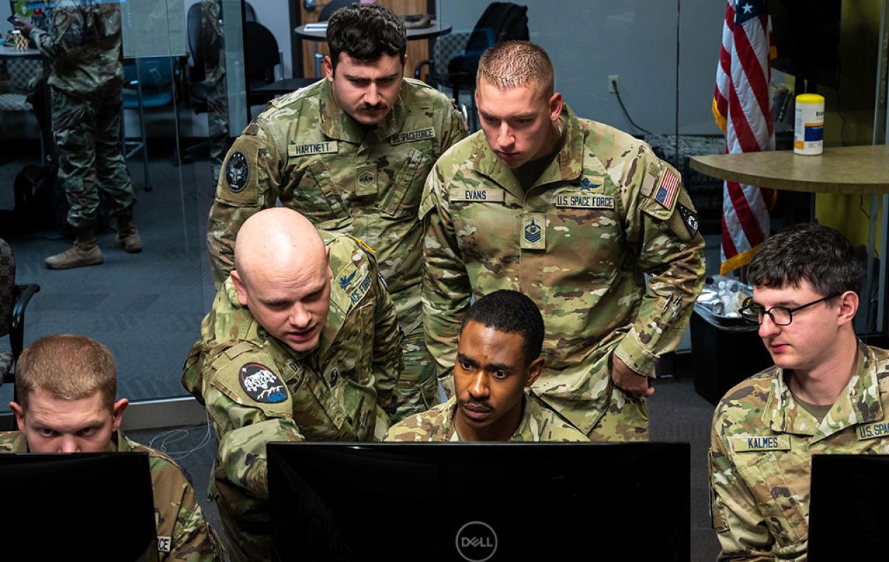 Military personnel, wearing camoflague uniform crowd round a computer monitor.