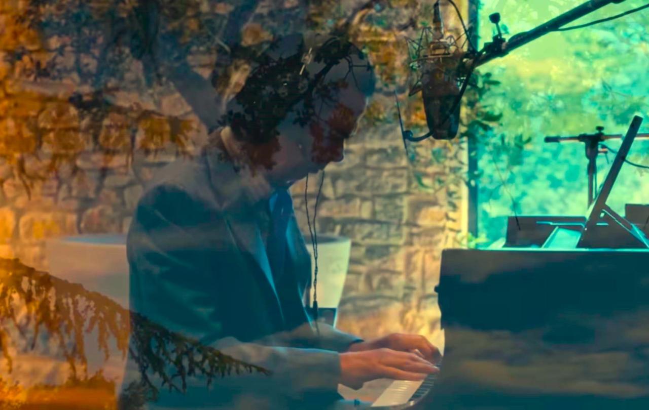 A pianist raises his arms while sitting at a grand piano amid recording equipment.