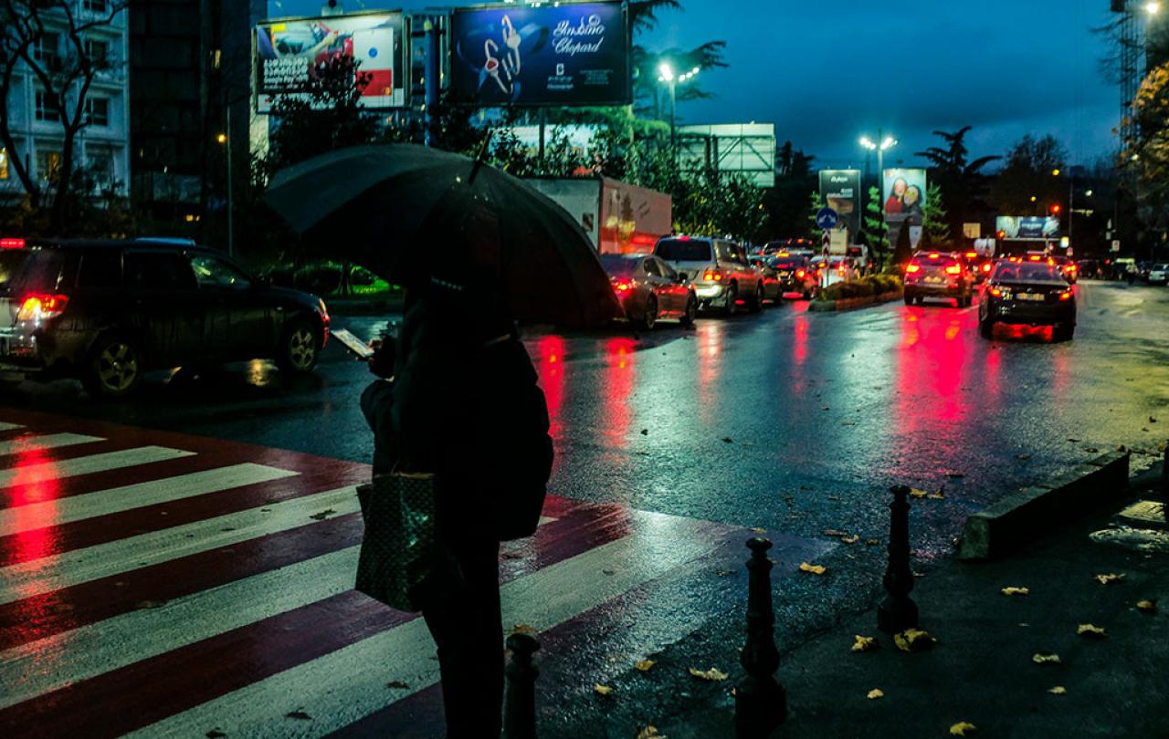 On a rainy night a pedestrian, holding a brolly, waits to cross a road.