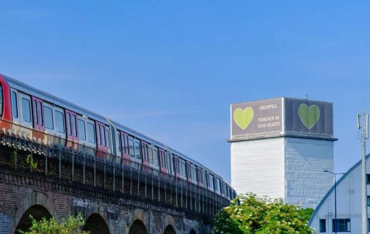 A tube train runs on a raised track, in the distance is a tower block wrapped in white material with a green heart on it.