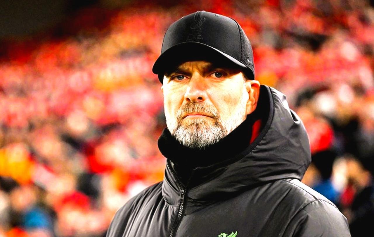 A stern looking  football manager stares hard, with a stand of supporters behind him.