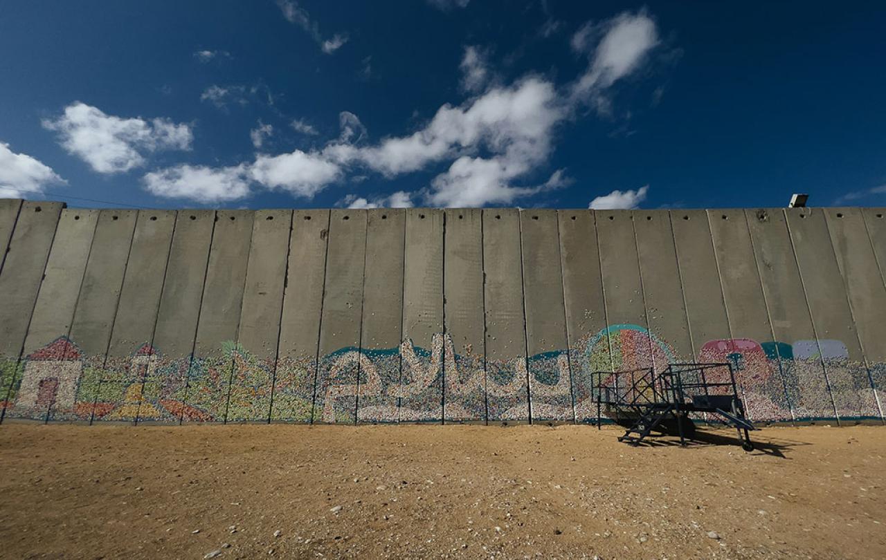 A graffitied concrete border wall stands below a blue sky and dusty ground