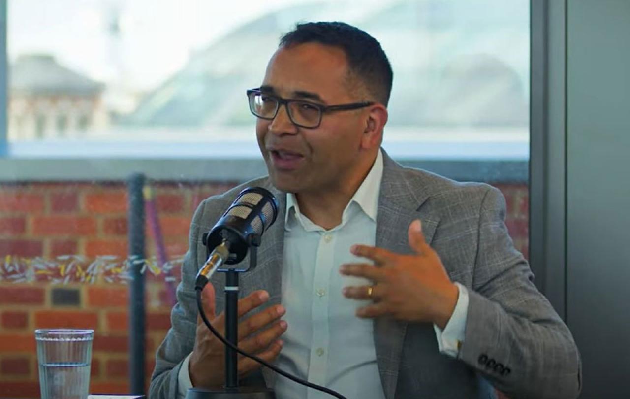 A man sitting at a table with a mic on it, raises his hands in gesture as he speaks. 