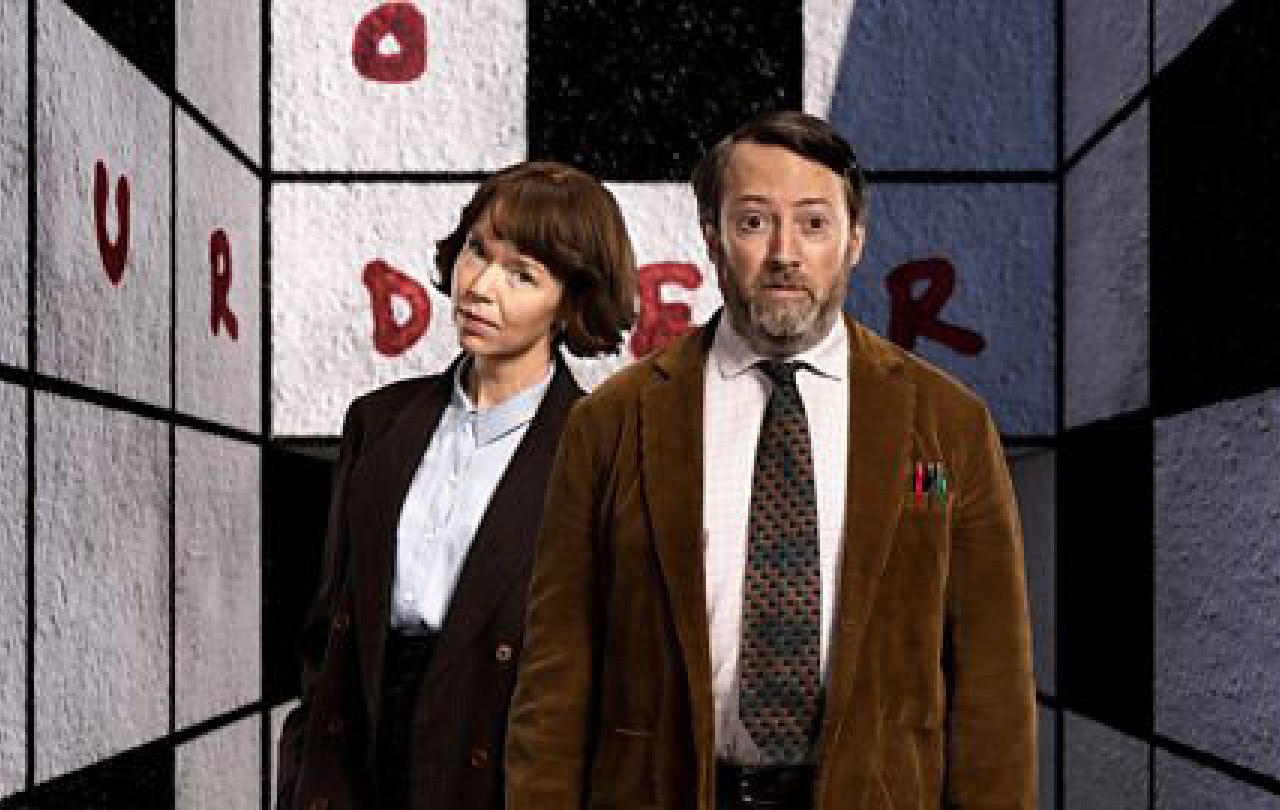 Two TV characters, a man and a woman, stand in front of a crossword cover walls of a room. 