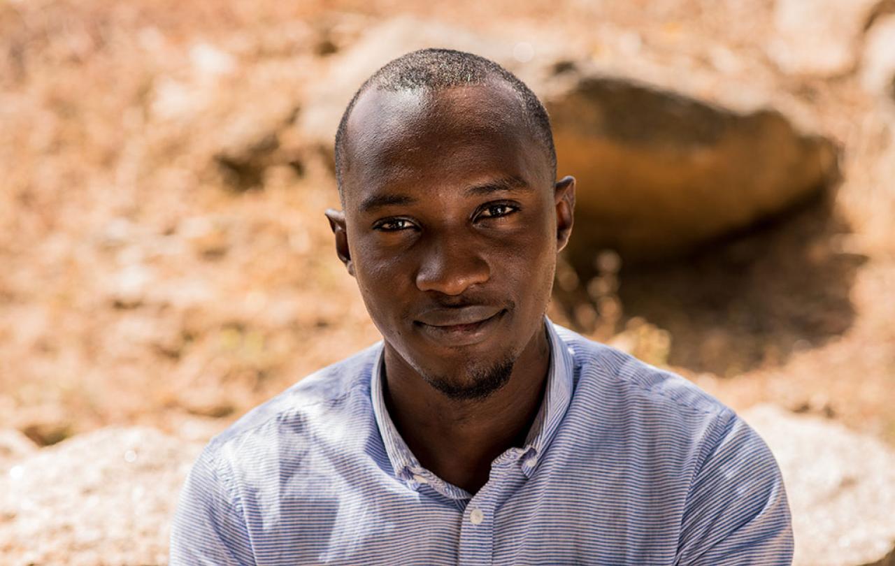 A Nigerian man looks up towards the camera, behind him is dusty ground