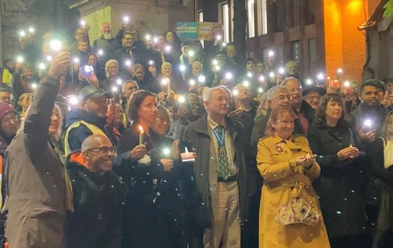 An outdoor vigil is lit by people holding up mobile phone lights.