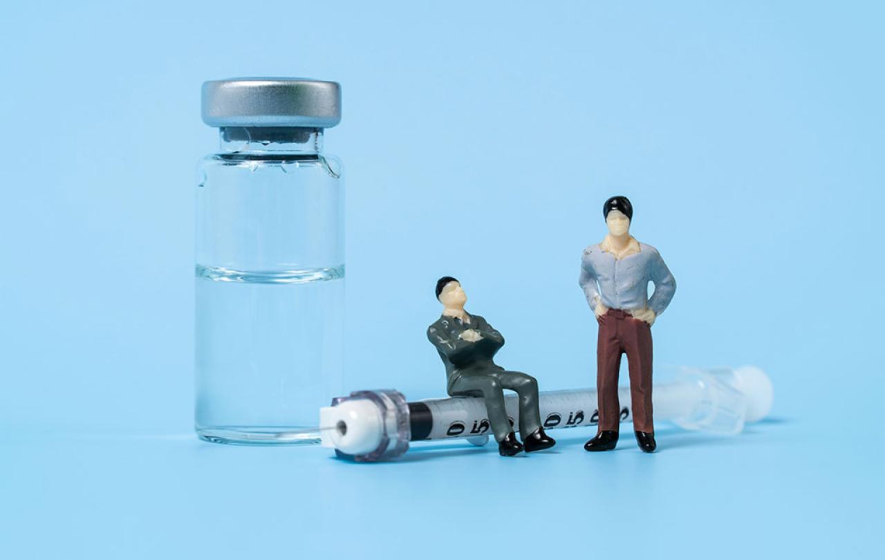 A tableau shows minature figures of two people, one sitting on a life size syringe and the other stands