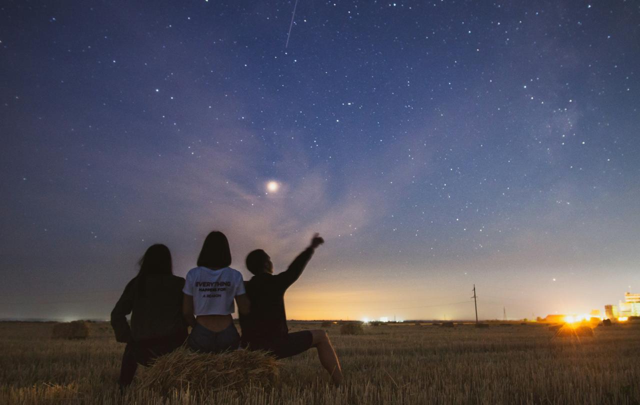 At dusk, three people sit on a field edge and look at the stars emerging.