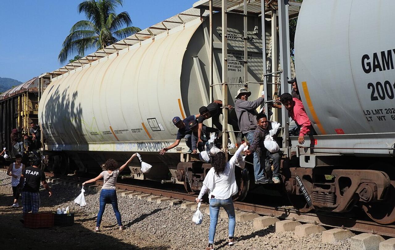 Migrants on a freight train reach for food bags held aloft by people on the track side.