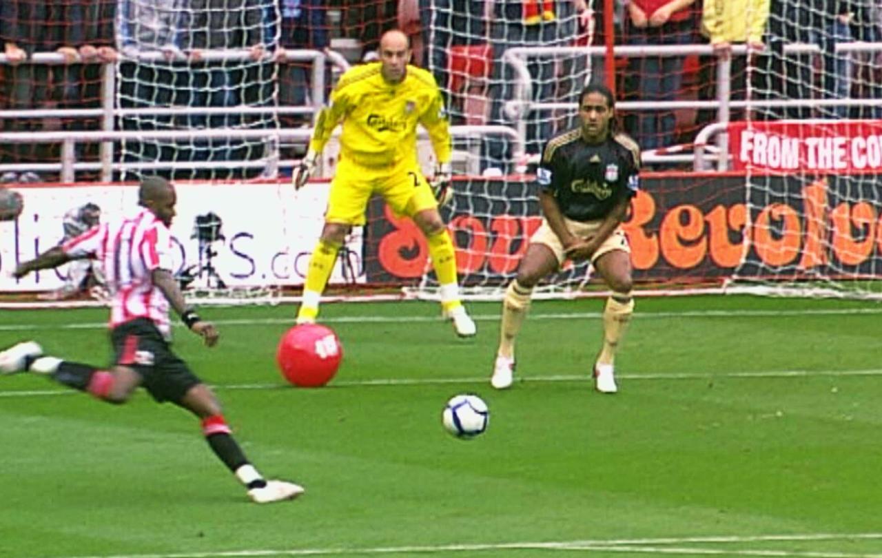 A striker is about kick a football towards a goal, a red beach ball sits between him and the goalkeeper.