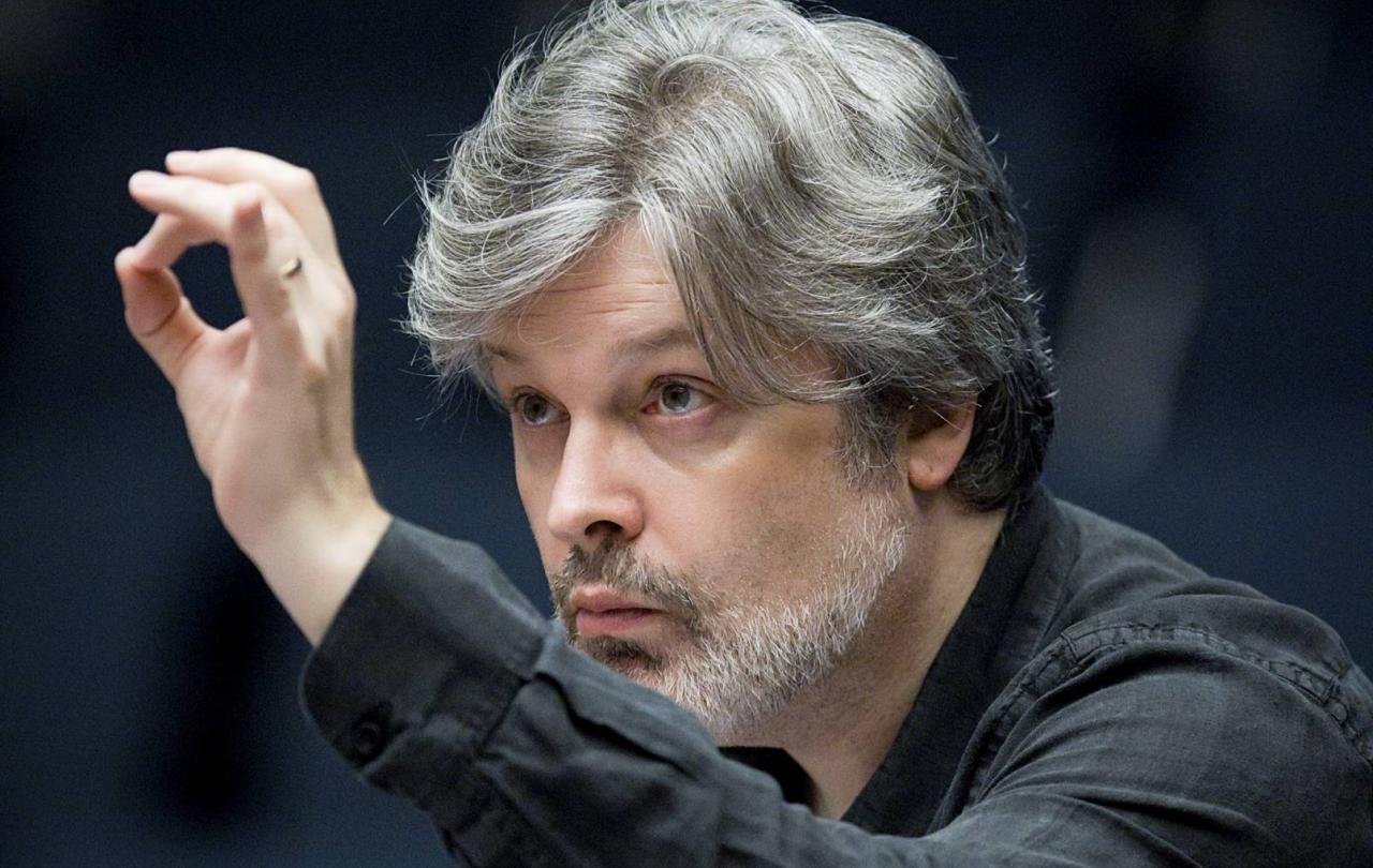 A conductor leans in toward an unseen orchestra with a raised hand.