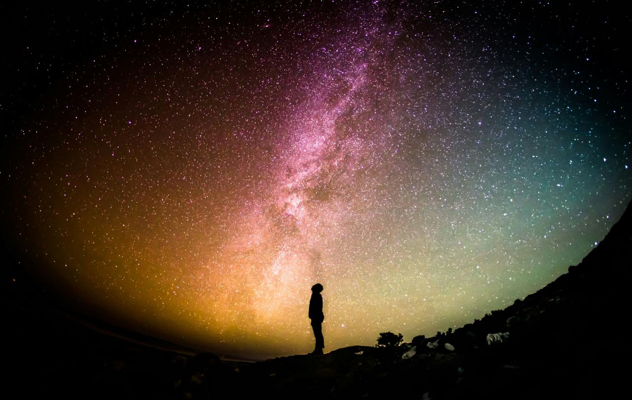 A fish eye lens view of a person standing silhouetted, looking up to a colourful night sky with the Milky Way across it