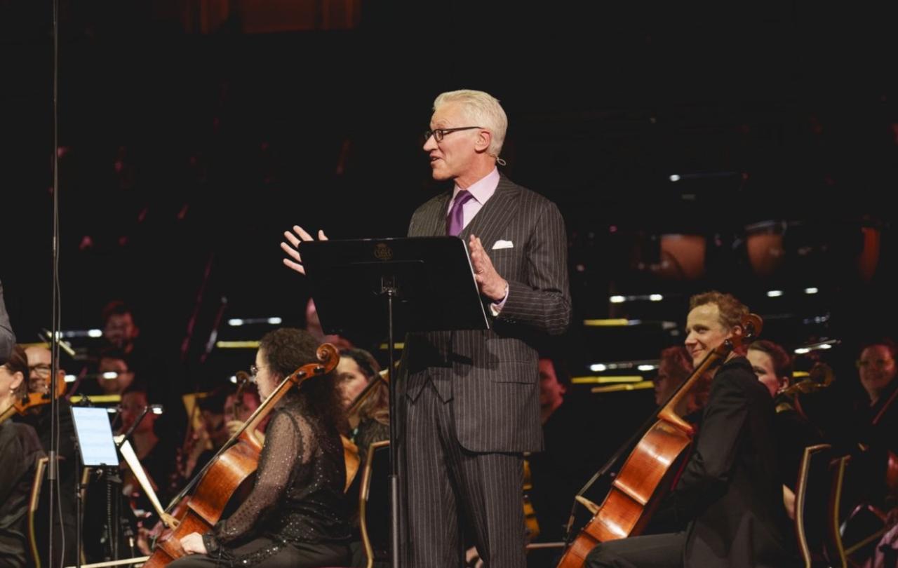 A man in a suit stands in front of a orchestra, by a lectern, gesturing while talking.