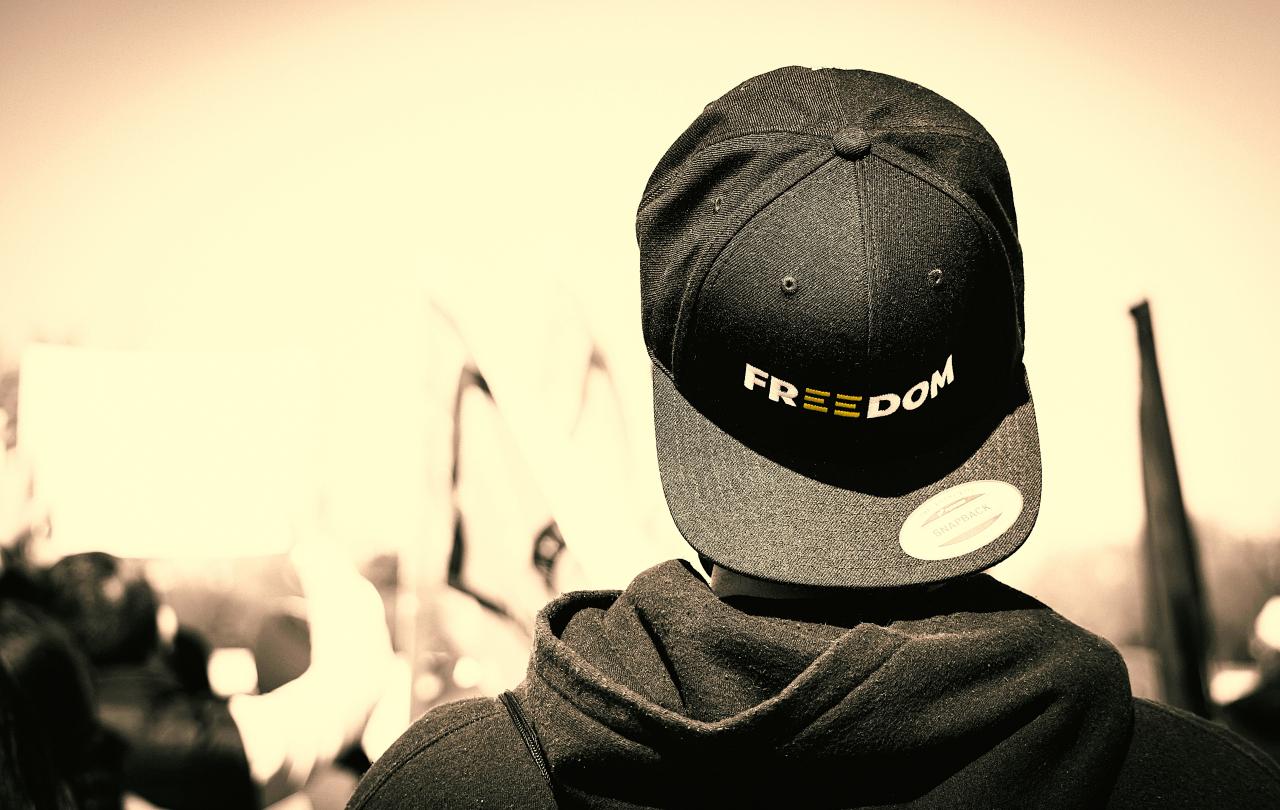 A protester stands with back to the camera, his baseball hat is turned backwards, it reads 'freedom'.